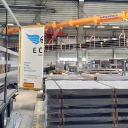 ECSI SPEDYCJA SPÓŁKA Z OGRANICZONĄ ODPOWIEDZIALNOŚCIĄ - Wnętrze hali magazynowej: suwnica transportuje metalowe płyty nad stosem zabezpieczonych folią arkuszy, w tle widoczny pracownik w szarym uniformie i wózek widłowy.