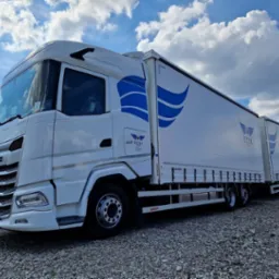 Biała ciężarówka DAF z naczepą i przyczepą, z niebieskim logo ECSI, zaparkowana na żwirowym placu pod błękitnym niebem z chmurami.