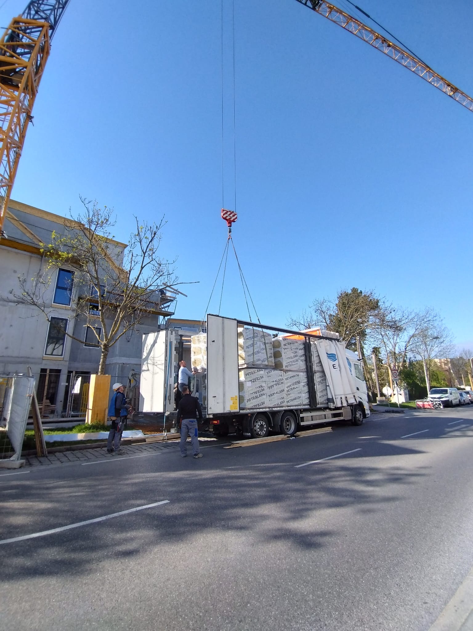 Rozładunek materiałów budowlanych z naczepy ciężarówki za pomocą dźwigu na tle budynku w trakcie budowy, widoczni pracownicy w kaskach.