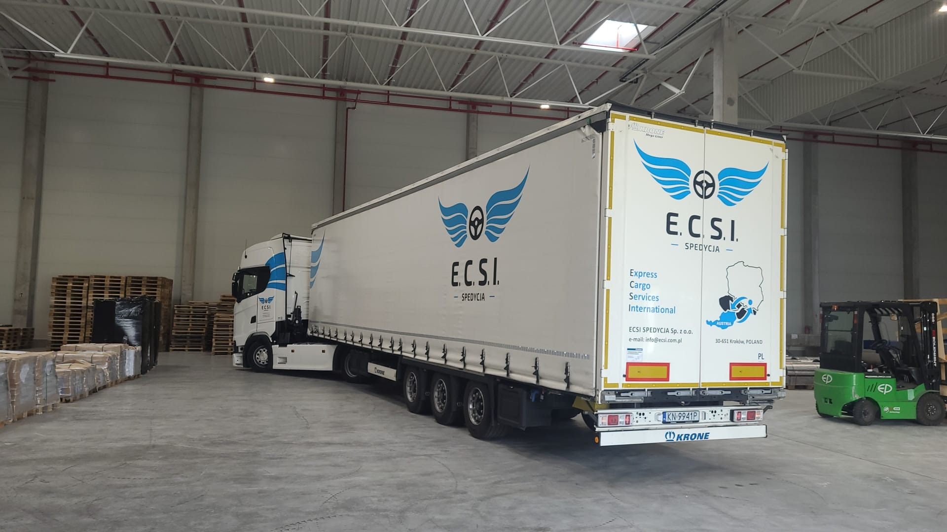 Ciężarówka z naczepą firmy ECSI Spedycja zaparkowana w magazynie, widoczny wózek widłowy i palety z towarem.