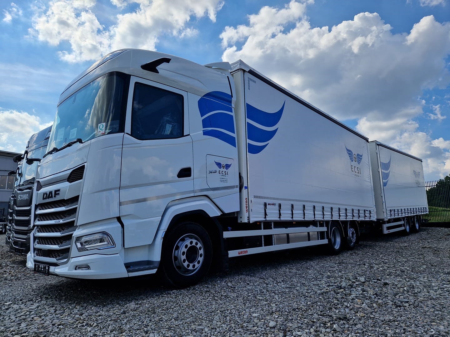 Biała ciężarówka DAF z naczepą i przyczepą, z niebieskim logo ECSI, zaparkowana na żwirowym placu pod błękitnym niebem z chmurami.
