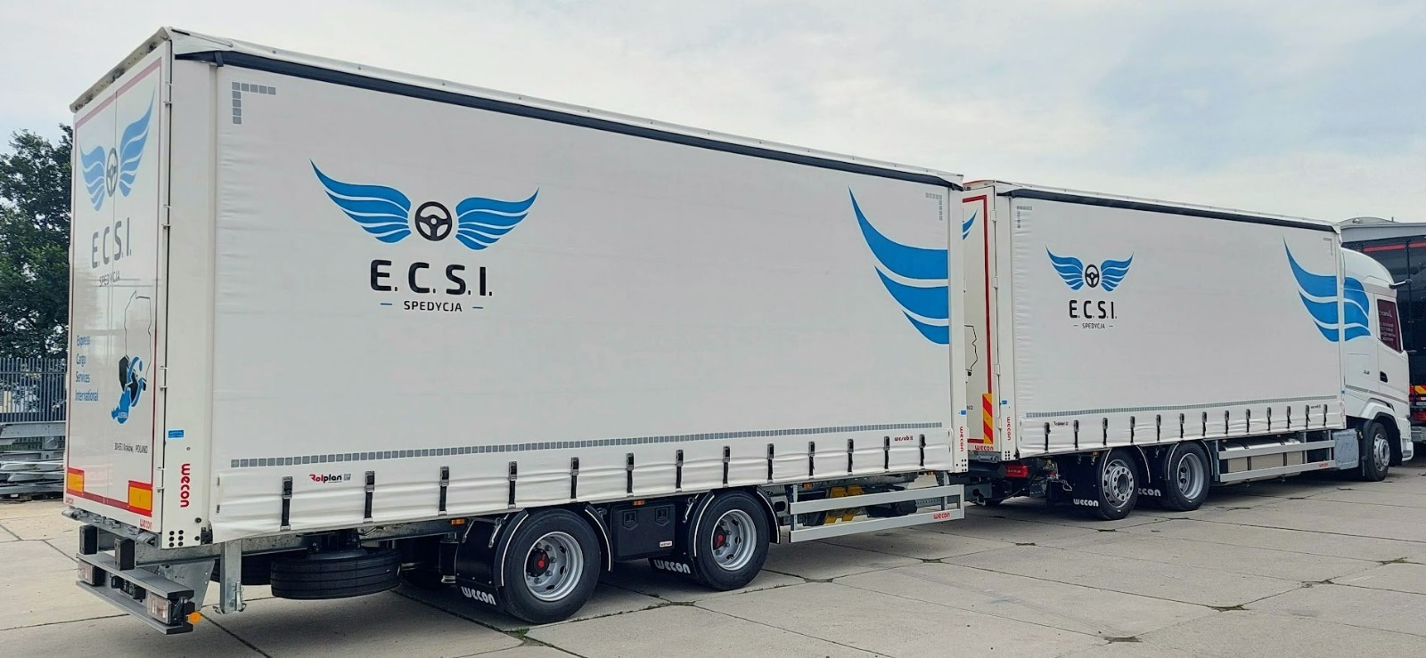Dwa białe naczepy ciężarowe z logo firmy spedycyjnej ECSI, zaparkowane na betonowym placu.