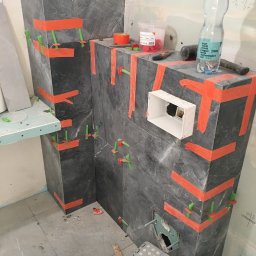 P&G Piotr Główczyński - Układanie ciemnych płytek imitujących kamień na ścianie w łazience, zabezpieczonych pomarańczową taśmą i zielonymi krzyżykami dystansowymi, widoczny otwór na gniazdko elektryczne, wiadro z zaprawą...