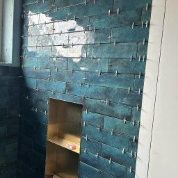 P&G Piotr Główczyński - Ściana łazienki wyłożona błyszczącymi, turkusowymi płytkami w układzie poziomym, z krzyżykami dystansowymi, wbudowana złota półka z kosmetykami i gąbką, widoczne fragmenty białej ściany.