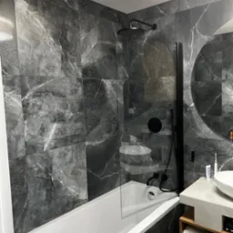 Nowoczesna łazienka z grafitowymi płytkami imitującymi marmur, białą wanną, czarną armaturą i okrągłym lustrem nad drewnianą szafką z ręcznikami.