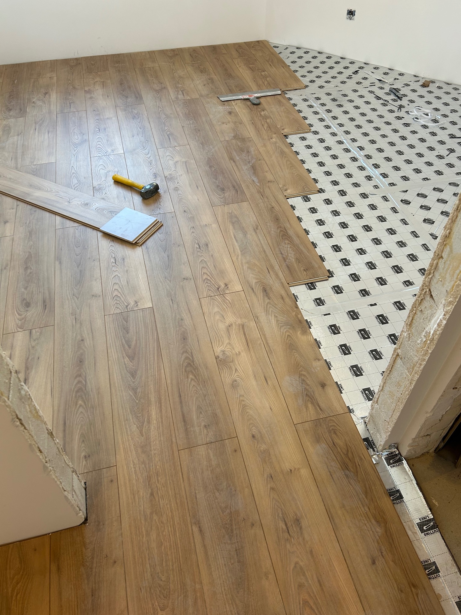 Układanie paneli podłogowych: widoczny fragment ułożonej podłogi, podkład z folią, młotek i miarka, w tle przygotowane podłoże z podkładem, surowy próg drzwiowy.