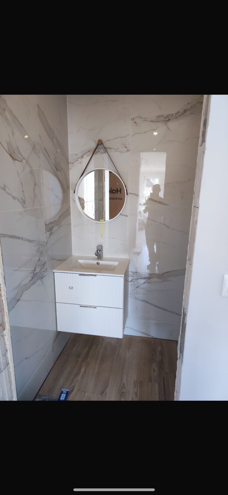 Nowoczesna łazienka z marmurowymi płytkami, okrągłym lustrem na skórzanym pasku i białą szafką pod umywalką, widoczny cień osoby fotografującej.