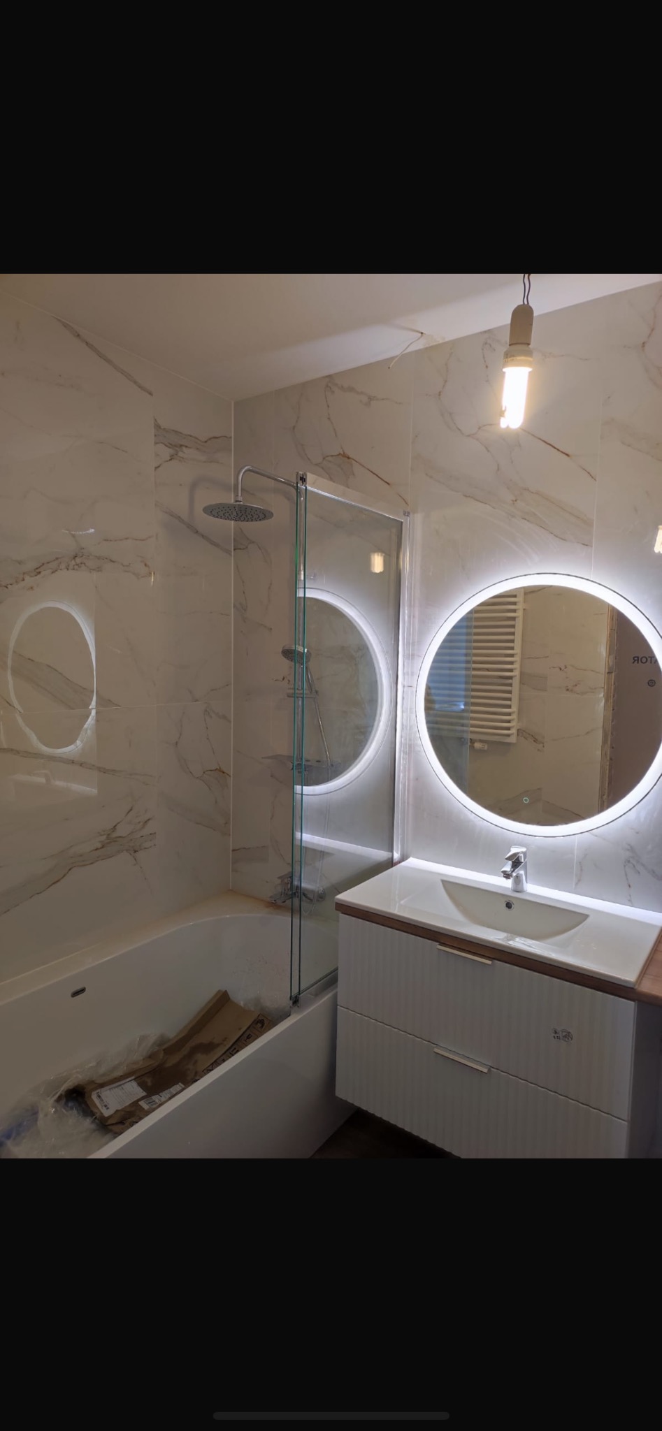 Nowoczesna łazienka po remoncie w Rumi z wanną z parawanem, okrągłym lustrem z podświetleniem LED i umywalką z szafką, wykończona płytkami imitującymi marmur.