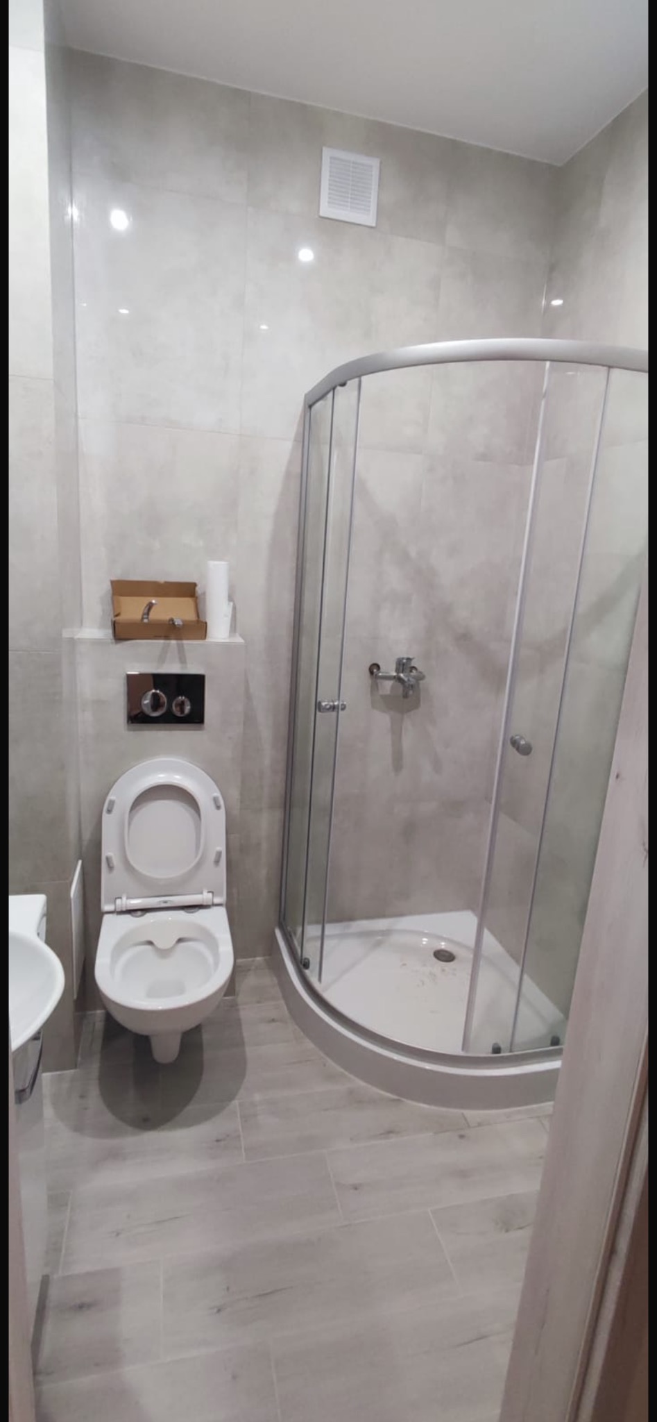 Nowoczesna łazienka w odcieniach szarości z kabiną prysznicową narożną, podwieszaną toaletą z chromowanym przyciskiem spłukującym oraz płytkami imitującymi drewno na podłodze. Na półce nad toaletą...