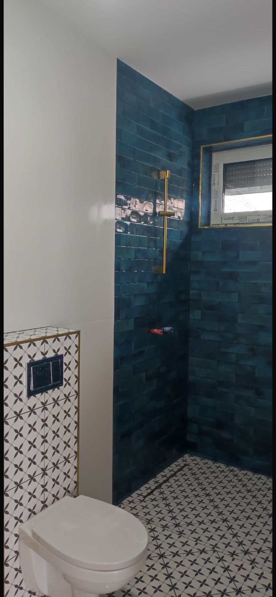 Nowoczesna łazienka z turkusową glazurą ułożoną w cegiełkę, złotą armaturą prysznicową i wzorzystą biało-czarną podłogą oraz zabudową WC z motywem kwiatowym.