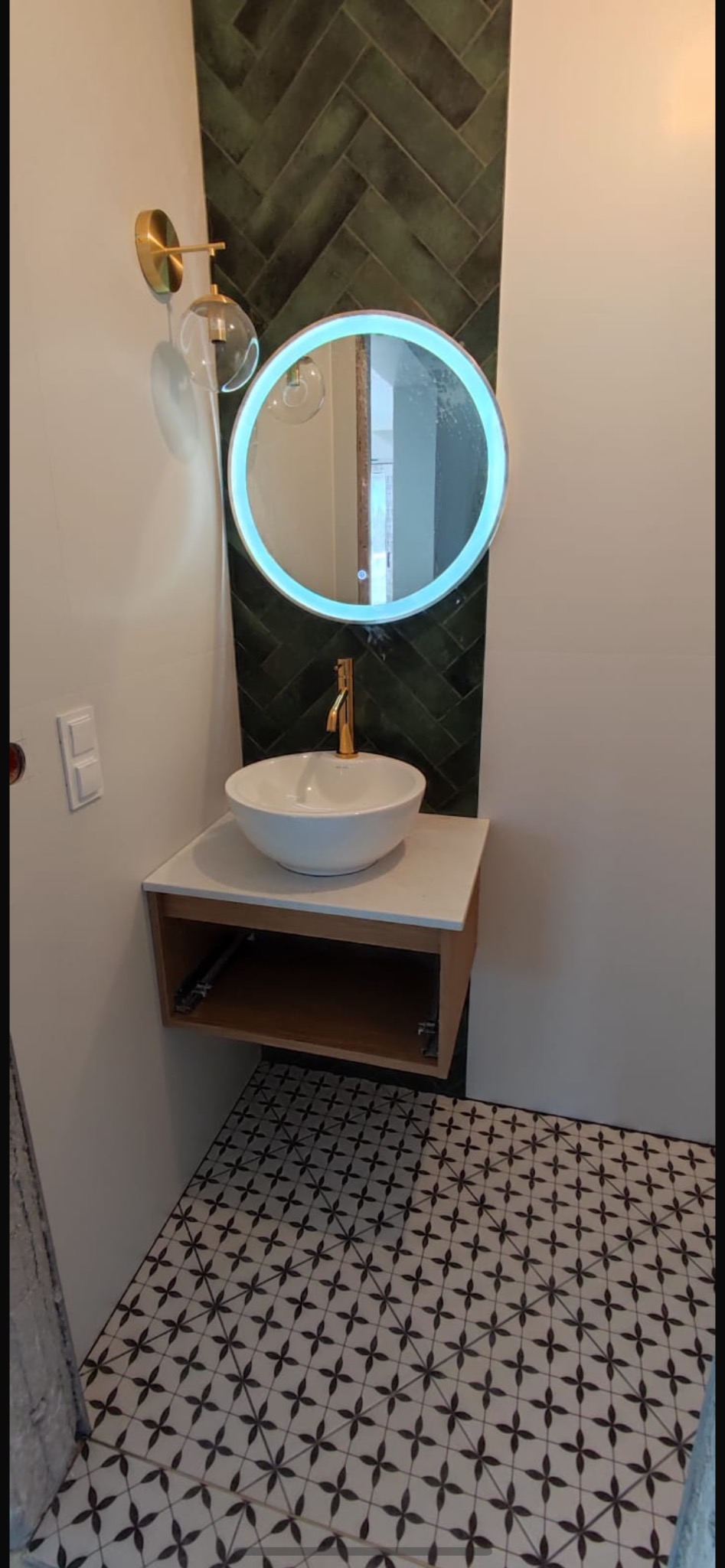 Nowoczesna łazienka z okrągłym lustrem podświetlanym LED, białą umywalką nablatową z złotą baterią, zielonymi płytkami ułożonymi w jodełkę i wzorzystą podłogą.