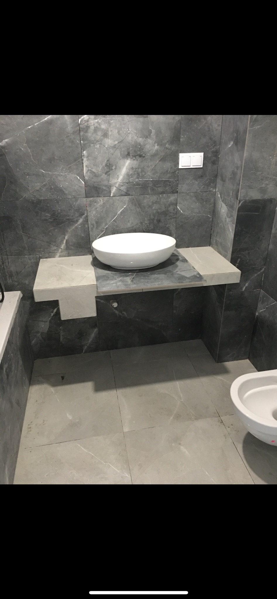 Nowoczesna łazienka z szarymi marmurowymi płytkami, okrągła biała umywalka nablatowa i częściowo widoczna toaleta.