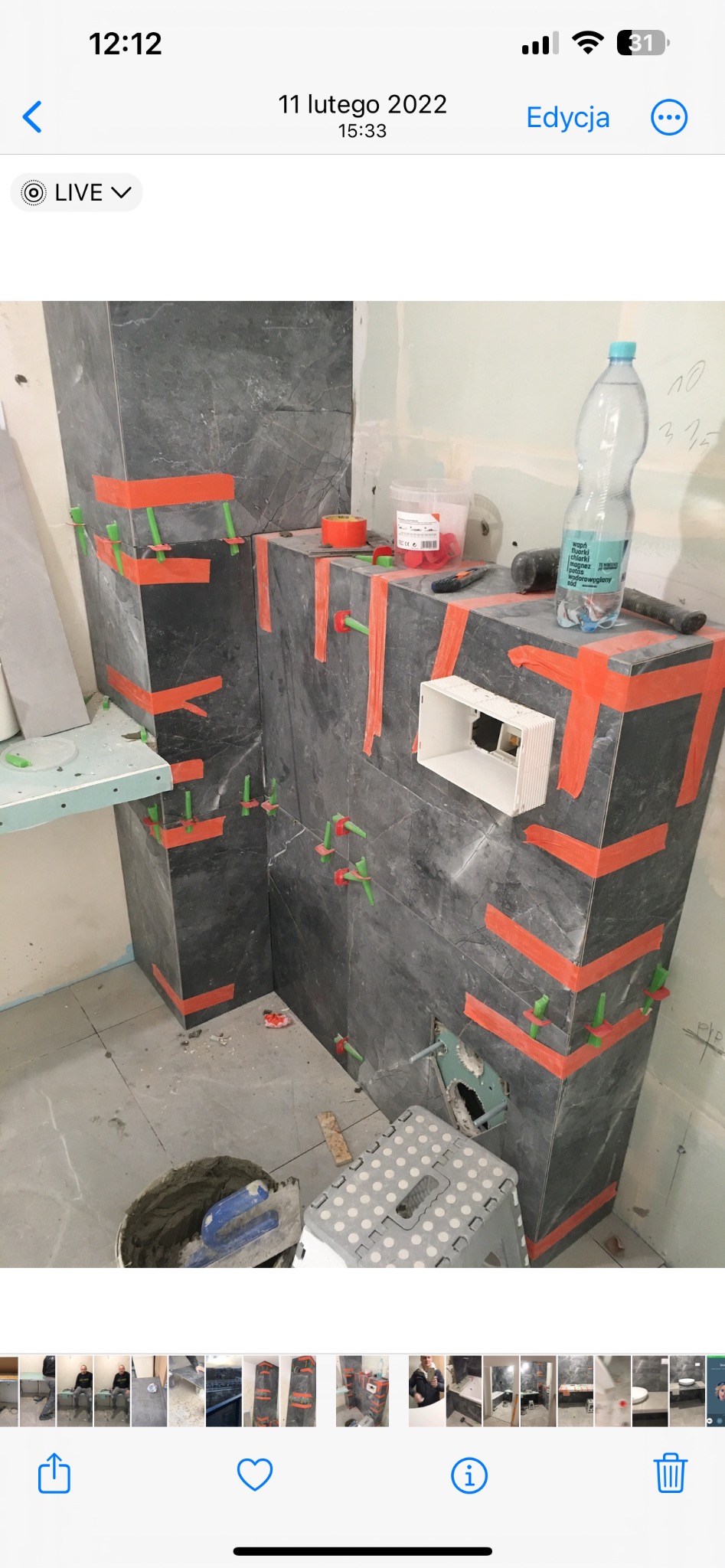 Układanie ciemnych płytek imitujących kamień na ścianie w łazience, zabezpieczonych pomarańczową taśmą i zielonymi krzyżykami dystansowymi, widoczny otwór na gniazdko elektryczne, wiadro z zaprawą...