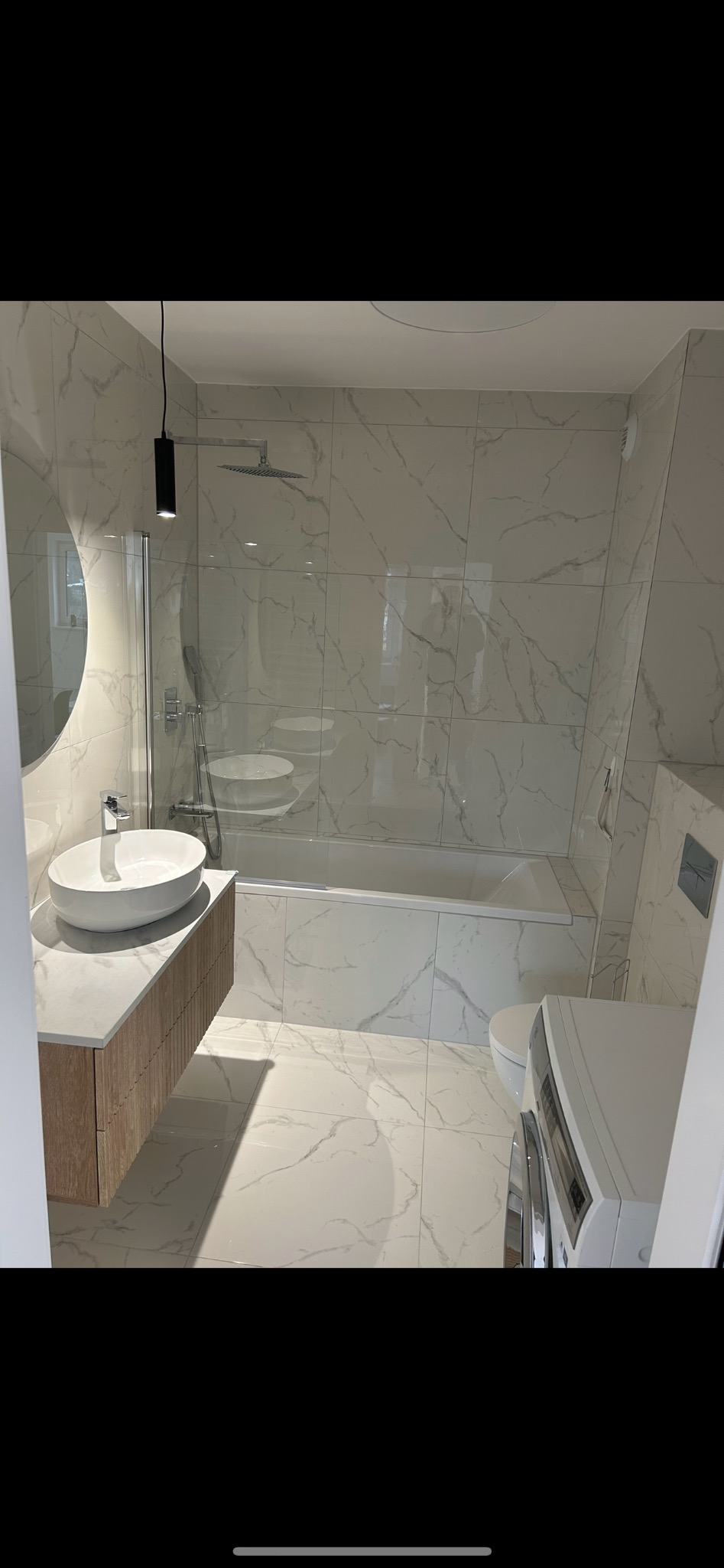 Nowoczesna łazienka z białymi marmurowymi płytkami, owalną umywalką na drewnianej szafce, czarną lampą wiszącą i szklanym panelem prysznicowym nad wanną.