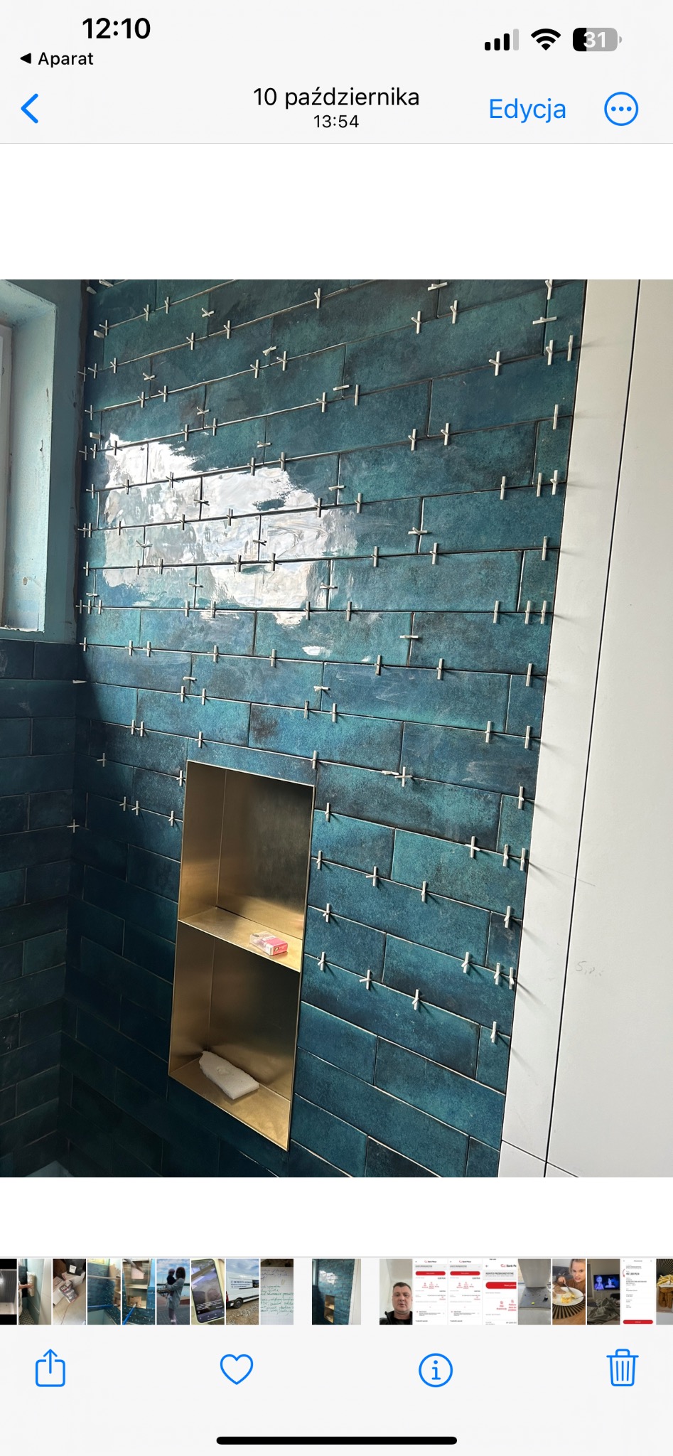 Ściana łazienki wyłożona błyszczącymi, turkusowymi płytkami w układzie poziomym, z krzyżykami dystansowymi, wbudowana złota półka z kosmetykami i gąbką, widoczne fragmenty białej ściany.