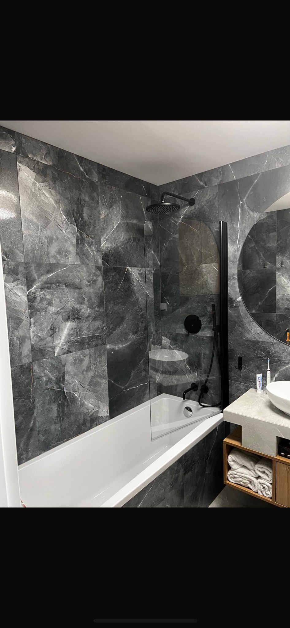 Nowoczesna łazienka z grafitowymi płytkami imitującymi marmur, białą wanną, czarną armaturą i okrągłym lustrem nad drewnianą szafką z ręcznikami.