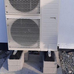 RaV Rafał Kaczor - Zewnętrzna jednostka pompy ciepła Panasonic na betonowych bloczkach, widoczna z bliska, z krzewem ozdobnym w tle.