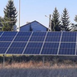 RaV Rafał Kaczor - Rząd paneli słonecznych zamontowany na metalowej konstrukcji w otoczeniu suchej trawy, w tle domy i znak STOP przy przejeździe kolejowym.