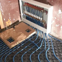 Rozdzielacz ogrzewania podłogowego z niebieskimi rurami ułożonymi na czarnej macie, widoczny karton z wyciętymi otworami, ściany w stanie surowym i butelka napoju na obudowie rozdzielacza.