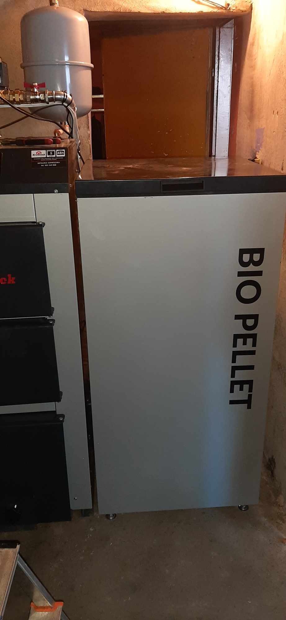 Szary piec na pellet BIO PELLET z widocznym zbiornikiem wyrównawczym i elementami instalacji w piwnicy z pomalowanymi ścianami.