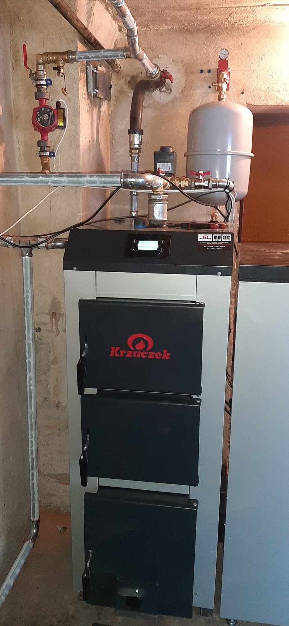 Zamontowany piec marki Krzaczek z widocznymi rurami, zaworami i zbiornikiem wyrównawczym w kotłowni.