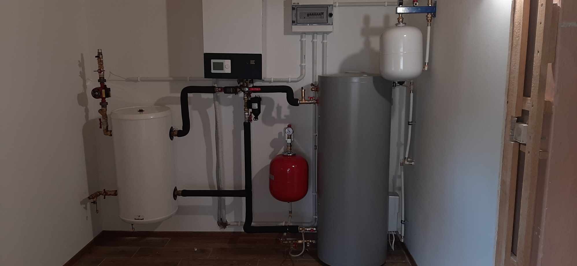 Zestawienie urządzeń grzewczych w kotłowni: dwa zbiorniki na wodę (biały i szary), czerwone naczynie przeponowe, czarna automatyka sterująca, instalacja hydrauliczna z rurami w izolacji.