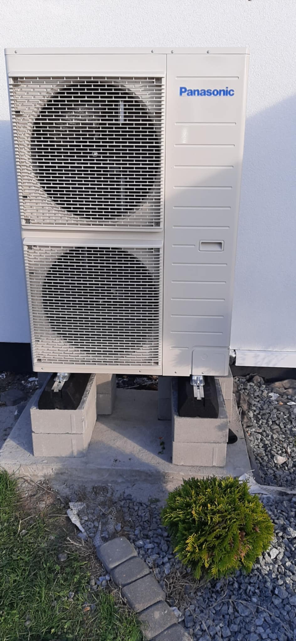 Zewnętrzna jednostka pompy ciepła Panasonic na betonowych bloczkach, widoczna z bliska, z krzewem ozdobnym w tle.