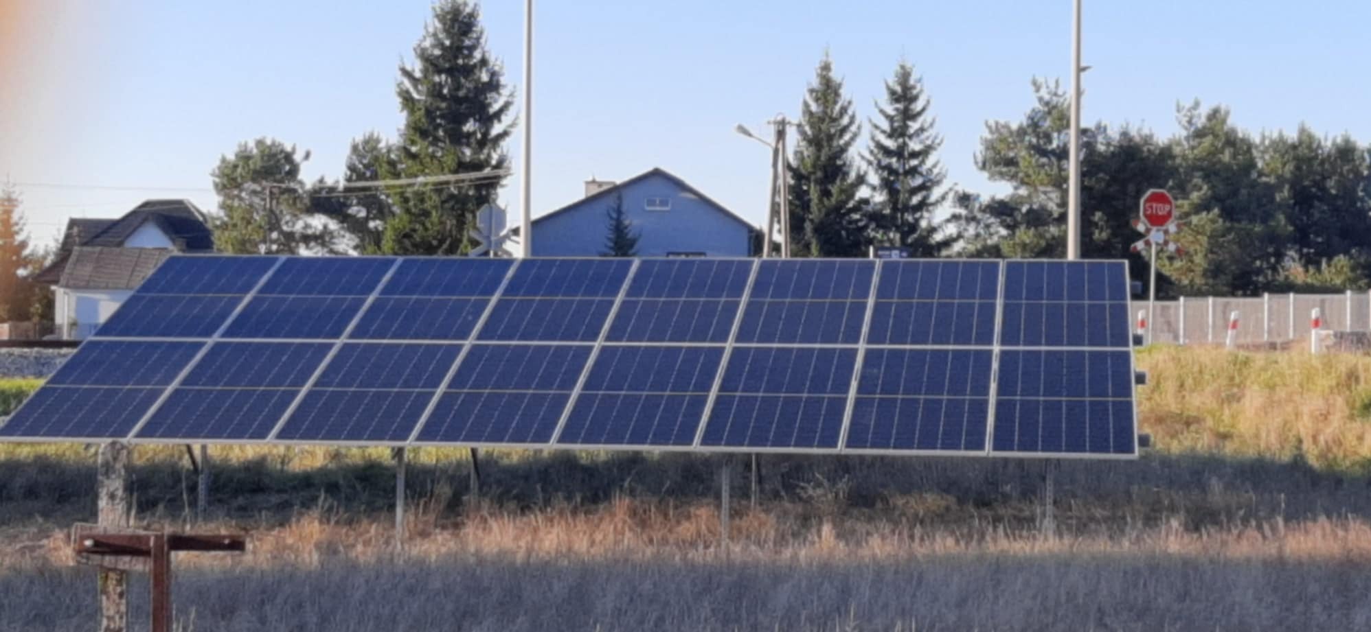 Rząd paneli słonecznych zamontowany na metalowej konstrukcji w otoczeniu suchej trawy, w tle domy i znak STOP przy przejeździe kolejowym.