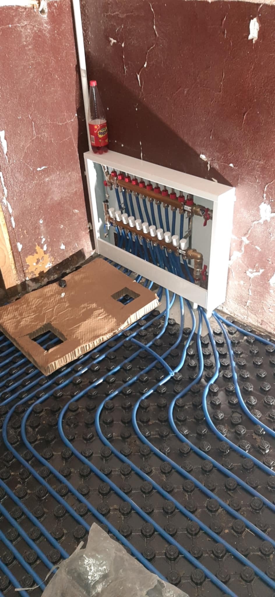 Rozdzielacz ogrzewania podłogowego z niebieskimi rurami ułożonymi na czarnej macie, widoczny karton z wyciętymi otworami, ściany w stanie surowym i butelka napoju na obudowie rozdzielacza.
