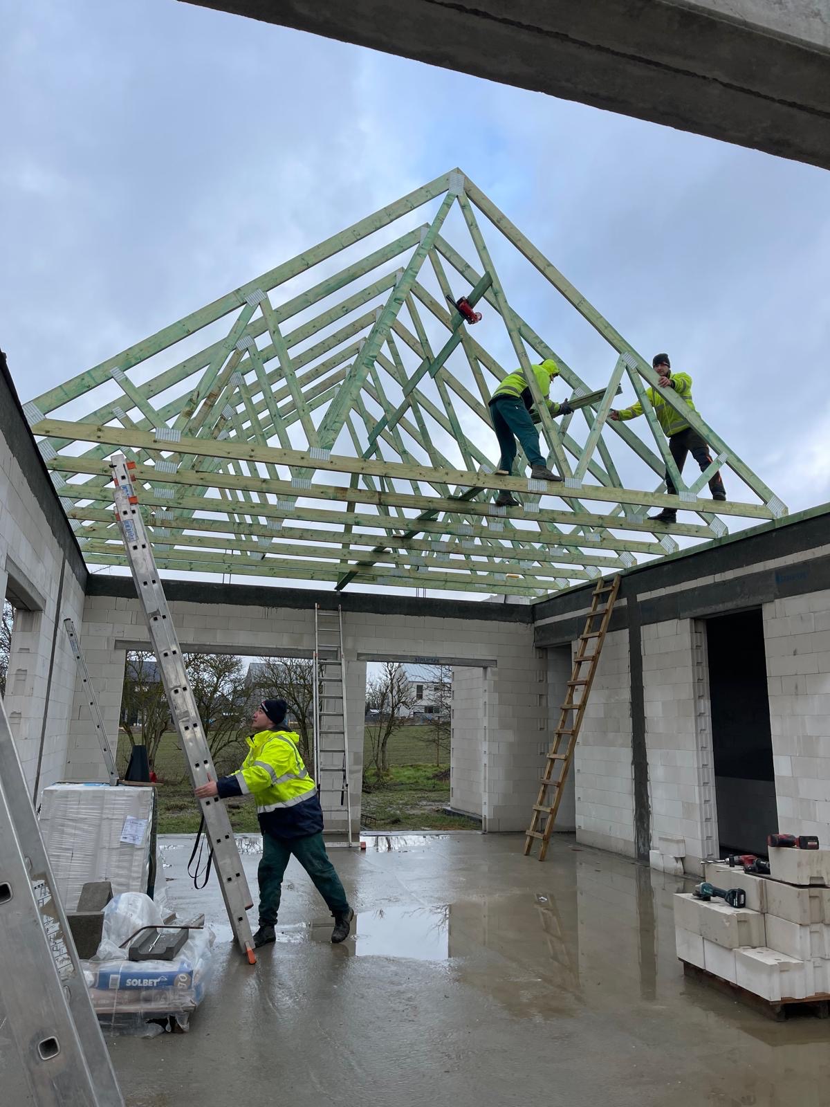 Montaż drewnianej konstrukcji dachu na surowym budynku z bloczków, widoczni trzej pracownicy w odblaskowych ubraniach, jeden używa piły elektrycznej, mokra posadzka wewnątrz budynku.