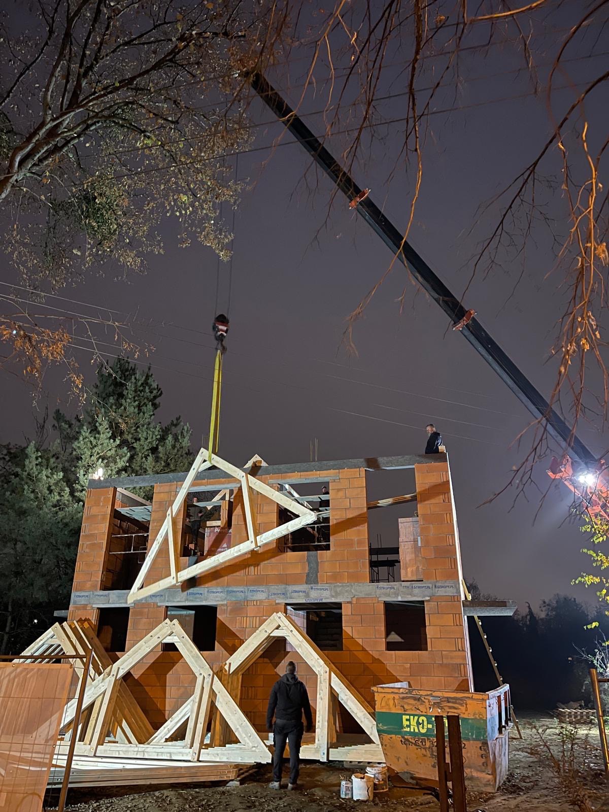 Montaż drewnianej więźby dachowej na murowanym budynku jednorodzinnym, widoczny dźwig i pracownicy, nocne ujęcie z dołu.