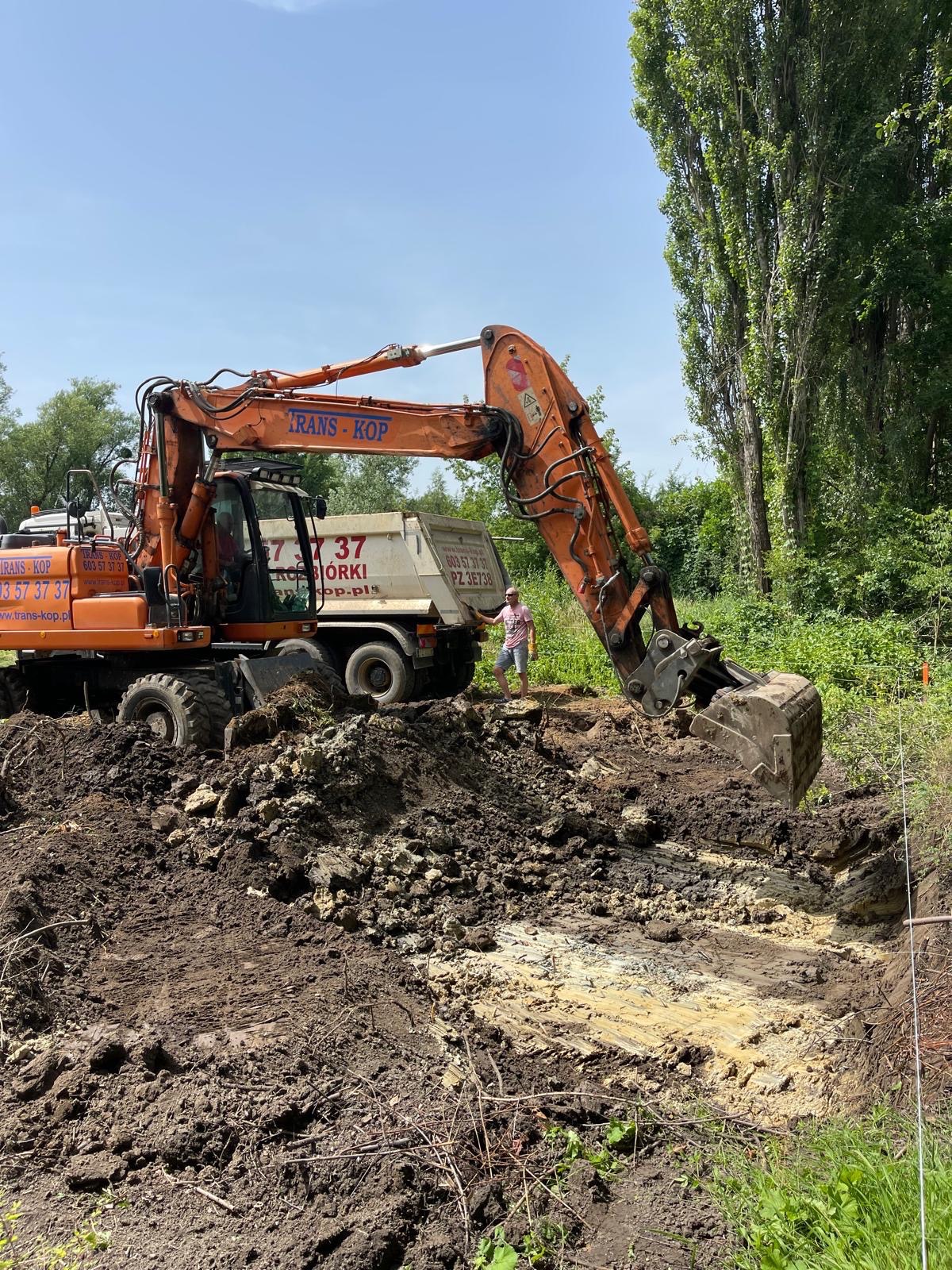 Pomarańczowa koparka kołowa Trans-Kop podczas prac ziemnych na placu budowy; widoczny wykop, usypana ziemia i pracownik w tle
