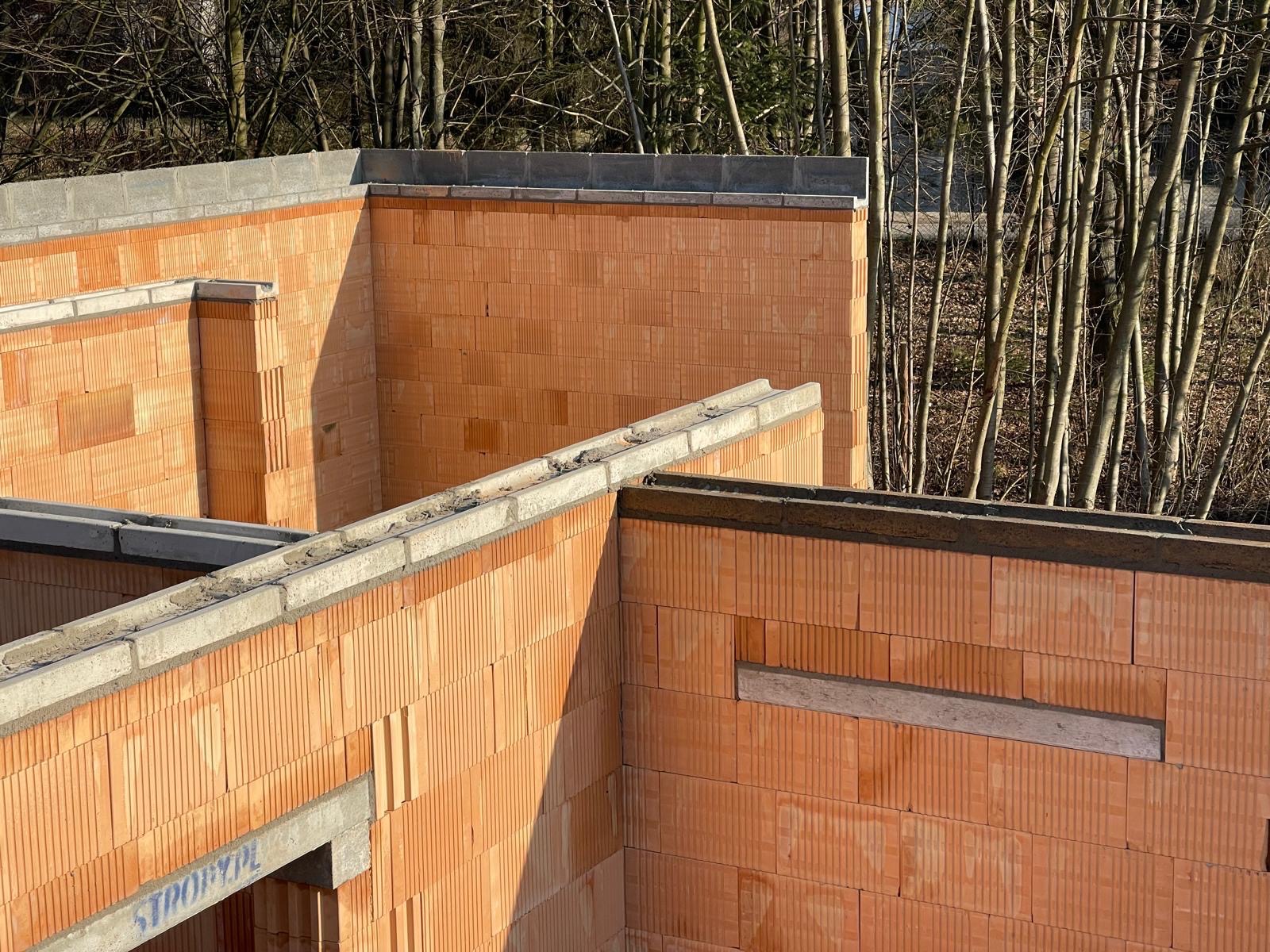 Wznoszone ściany budynku z czerwonej cegły ceramicznej z widocznymi elementami betonowymi, w tle drzewa bez liści w słoneczny dzień.