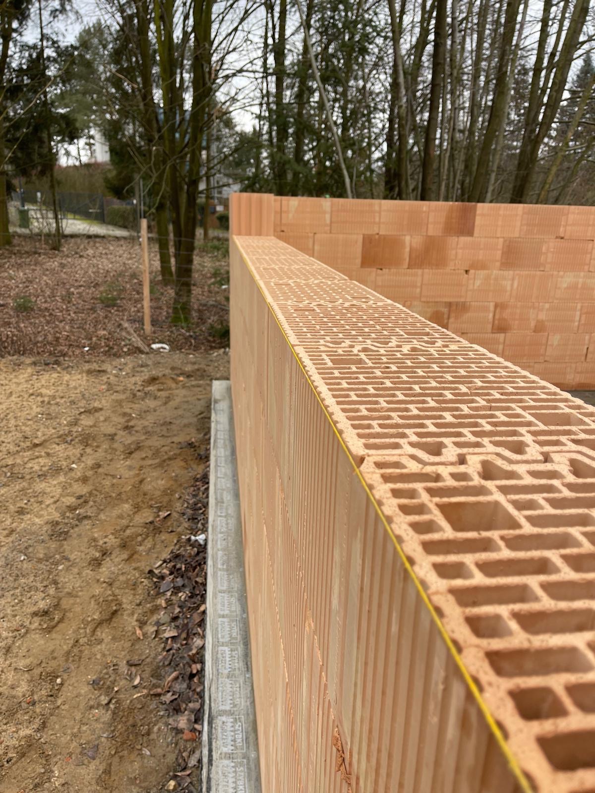 Wznoszenie muru z pustaków ceramicznych Porotherm z widoczną żółtą linką poziomującą i izolacją fundamentu, na tle drzew bez liści.