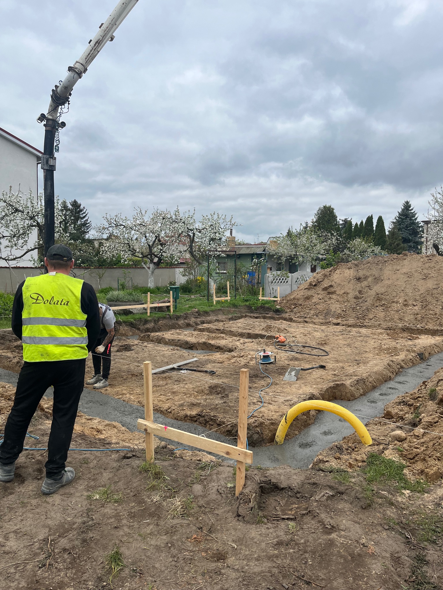 Wylewanie fundamentów pod budowę domu jednorodzinnego; widoczny pracownik w kamizelce z logo firmy, beton w wykopach, żółta rura wentylacyjna i ramię pompy do betonu.