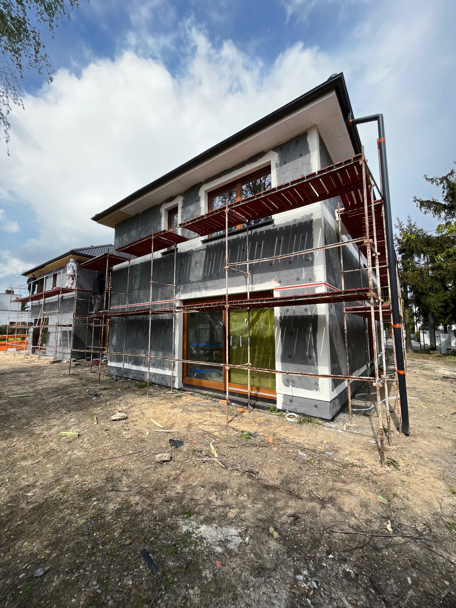 Nowoczesny dom jednorodzinny w trakcie ocieplania, widoczne rusztowania i ekipa budowlana w kombinezonach ochronnych, teren wokół domu częściowo zagospodarowany.