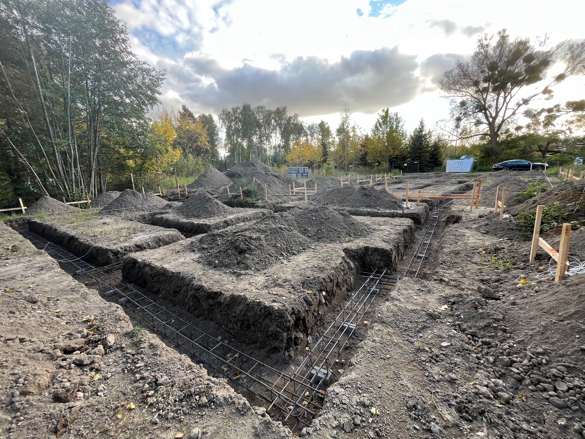 Wykopy pod fundamenty z ułożonym zbrojeniem, widoczne drewniane szalunki i hałdy ziemi na działce budowlanej, w tle drzewa i błękitne niebo z chmurami.