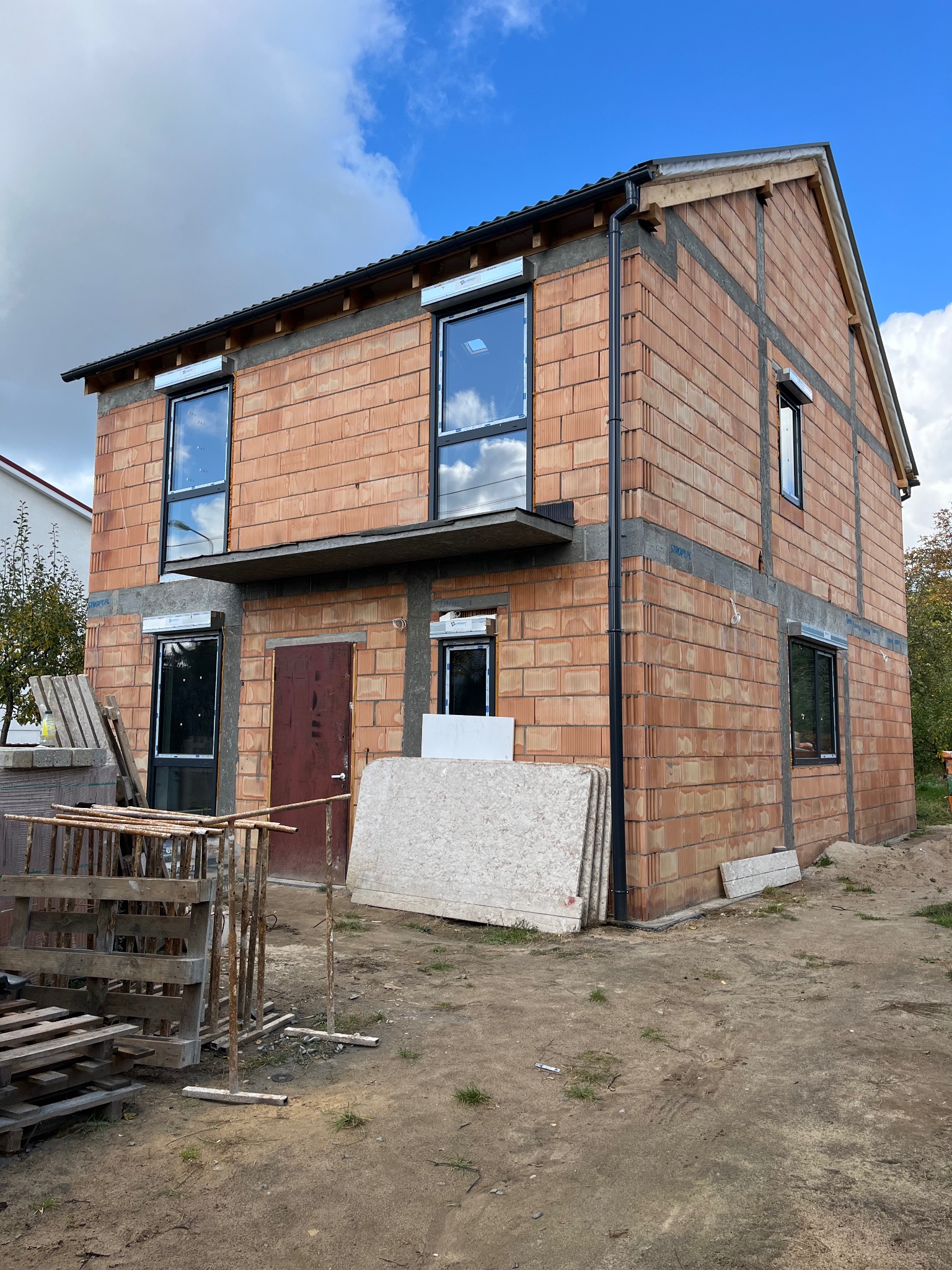 Dwukondygnacyjny dom w trakcie budowy z czerwonej cegły, z zamontowanymi oknami i przygotowanym miejscem na balkon, widoczne rusztowania i materiały budowlane.