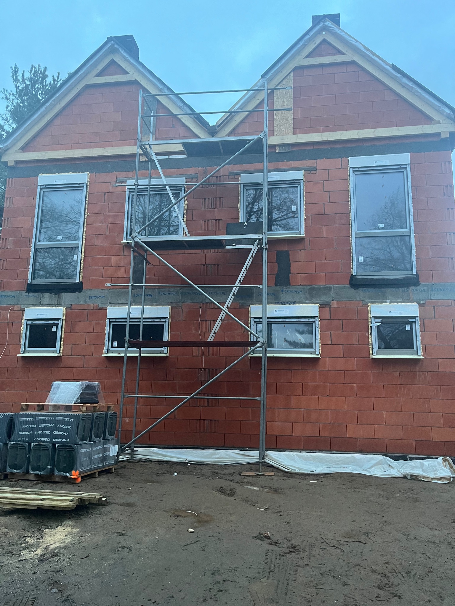 Wznoszenie dwukondygnacyjnego budynku z czerwonej cegły ceramicznej z zamontowanymi oknami; widoczne rusztowanie, palety cegieł i błotniste podłoże.