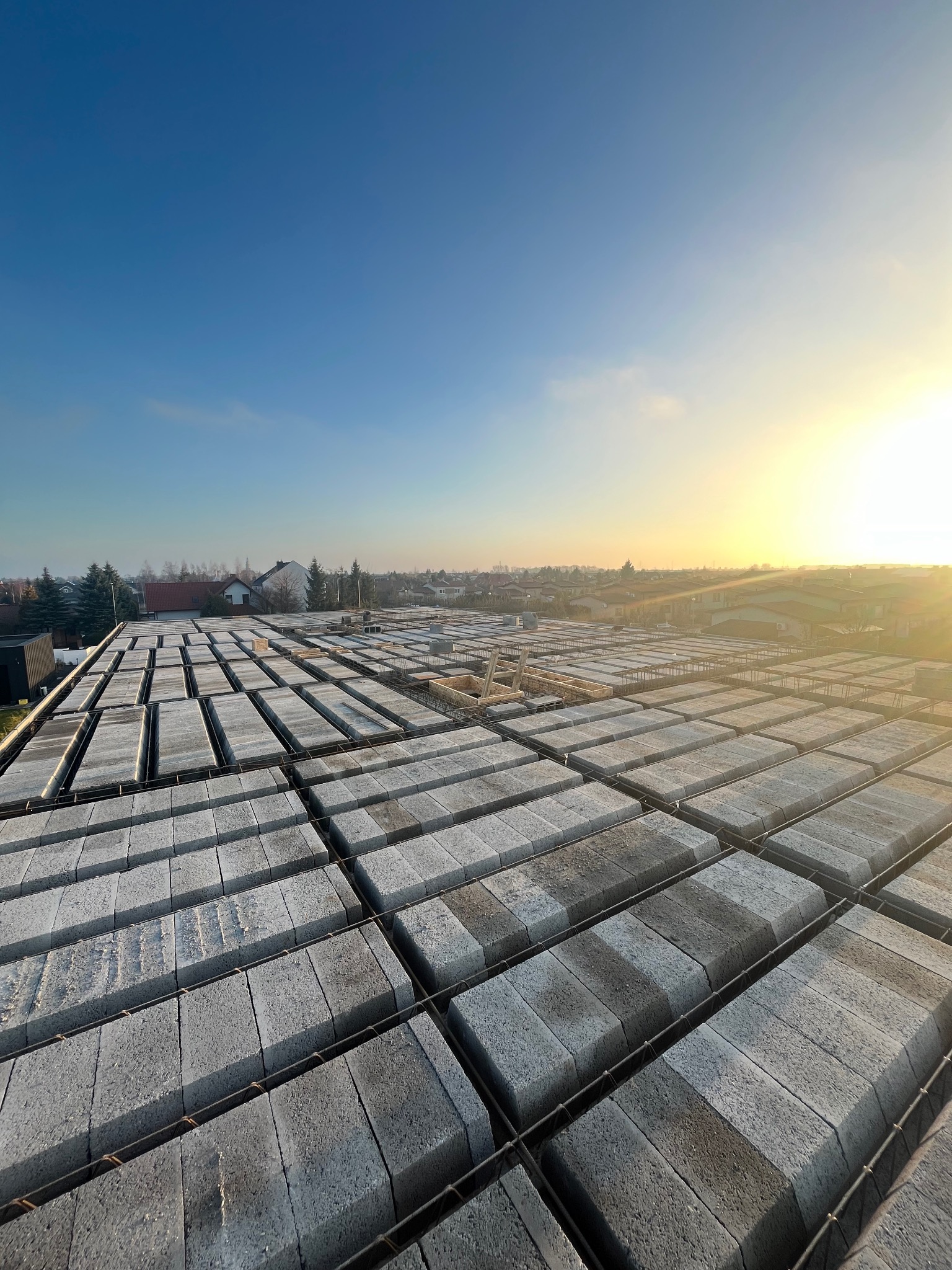 Wieniec stropowy z szalunkiem traconym wypełnionym bloczkami betonowymi, widok z góry na tle zabudowy jednorodzinnej w promieniach wschodzącego słońca.