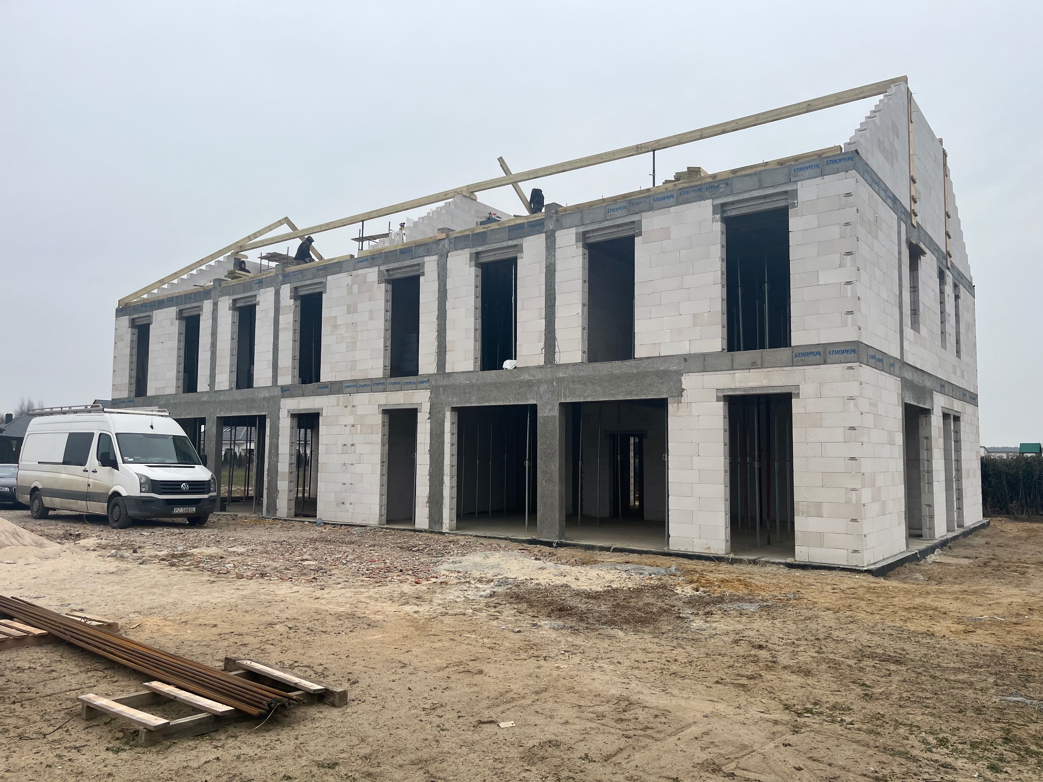 Wznoszenie dwukondygnacyjnego budynku z białych bloczków, widoczni pracownicy na dachu montujący drewnianą konstrukcję więźby, obok zaparkowany biały bus.