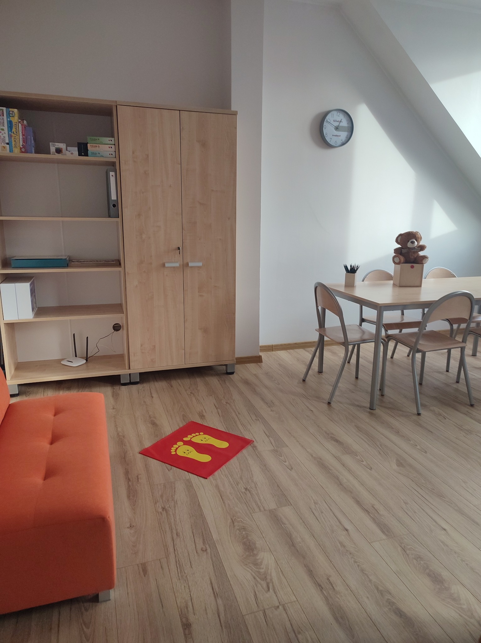 Jasne wnętrze pokoju z drewnianą podłogą, pomarańczową sofą, biurkiem z krzesłami i pluszowym misiem, szafą na książki i czerwoną matą z żółtymi stopami na podłodze.