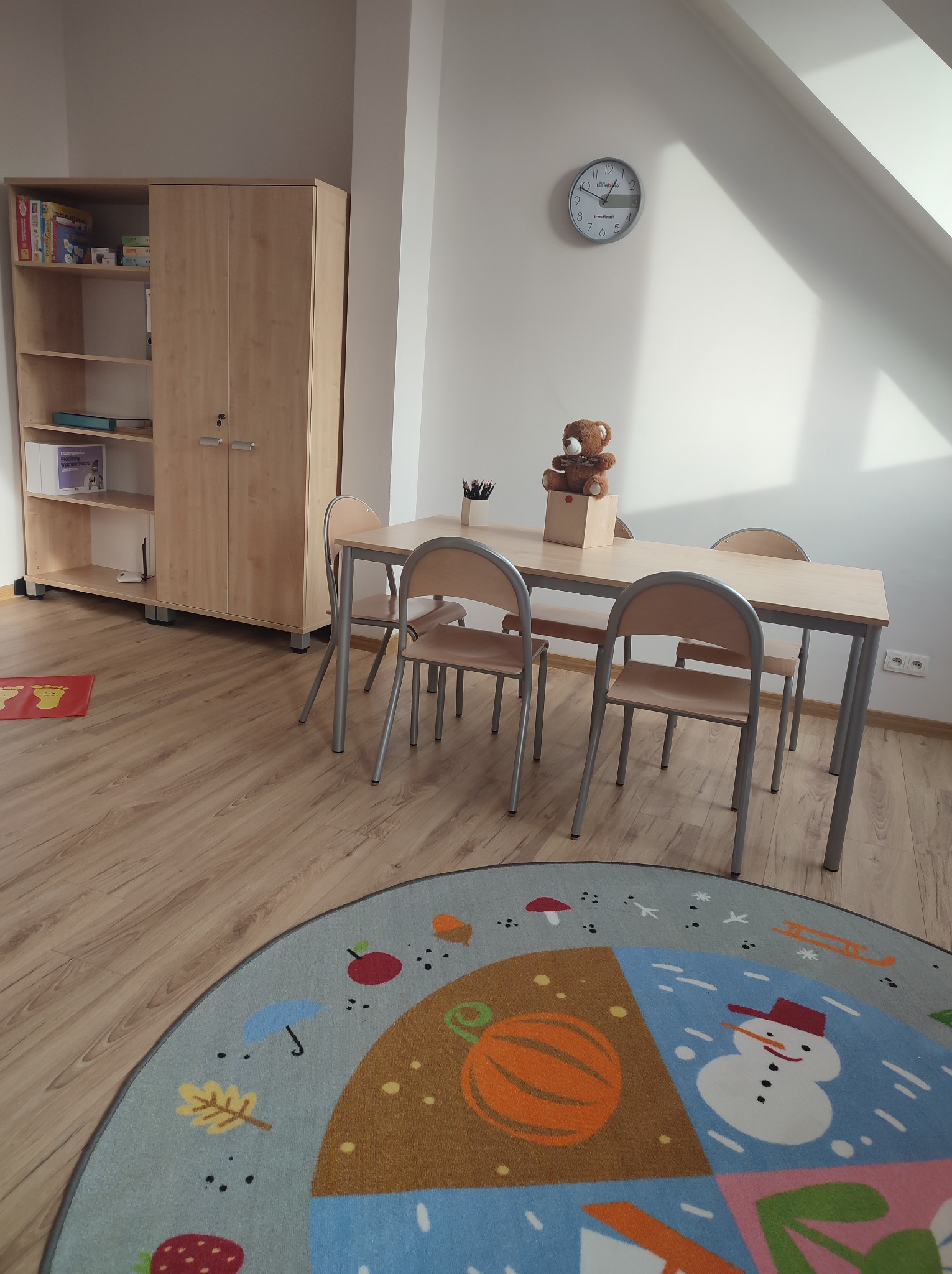 Jasne wnętrze pokoju zabaw z drewnianą podłogą, stolikiem z krzesłami, szafką na książki i okrągłym dywanem z kolorowymi wzorami, na stole stoi pluszowy miś w pudełku.
