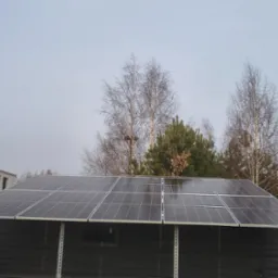 Gotowa instalacja paneli słonecznych na dachu budynku gospodarczego w pochmurny dzień. W tle drzewa i budynek w trakcie budowy.