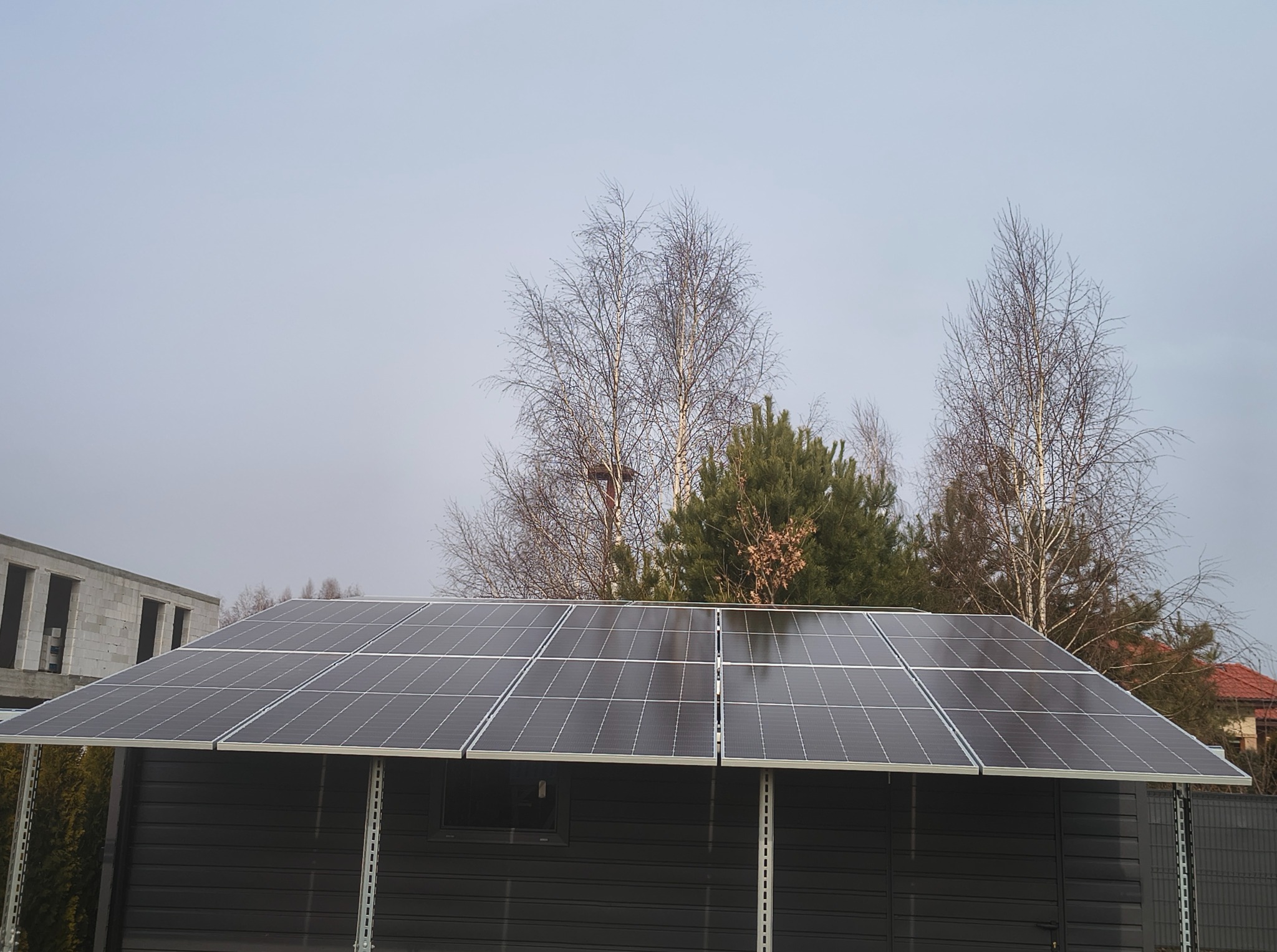 Gotowa instalacja paneli słonecznych na dachu budynku gospodarczego w pochmurny dzień. W tle drzewa i budynek w trakcie budowy.