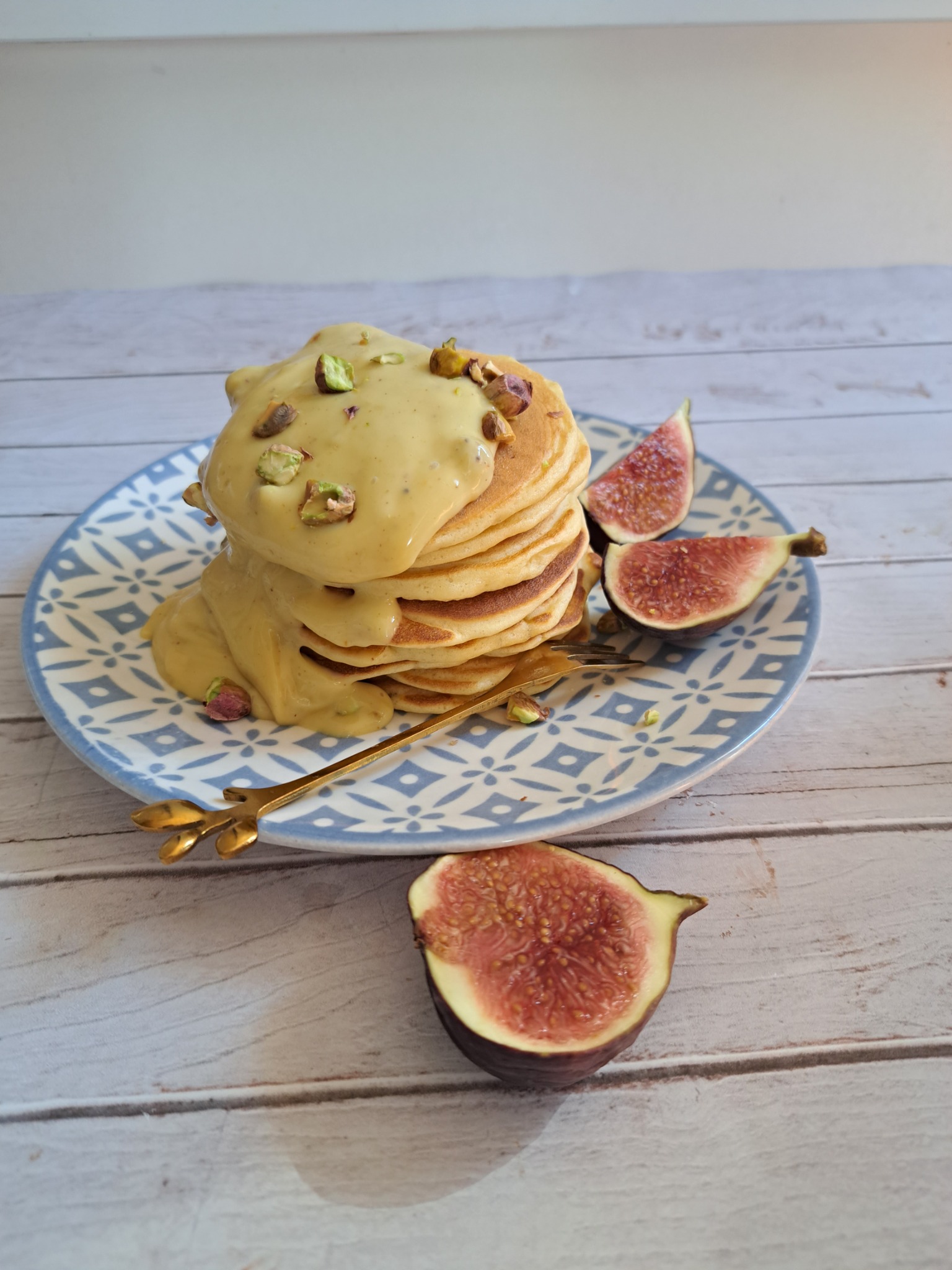 Stos pankejków polanych kremem pistacjowym i posypanych pistacjami, podane z połówkami fig na talerzu w niebieskie wzory, ze złotym widelcem na drewnianym stole.