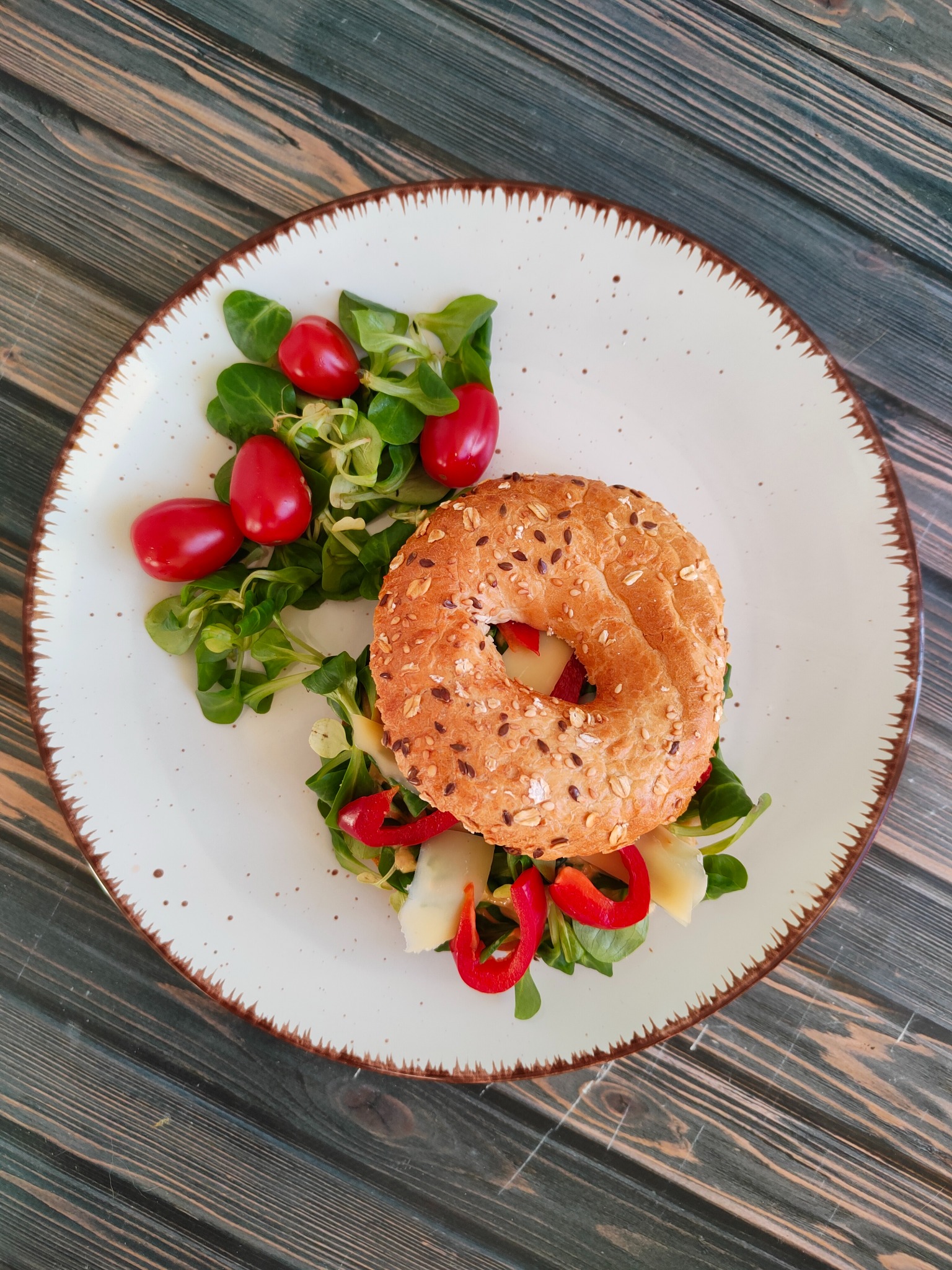 Bagel z ziarnami, serem i papryką na białym talerzu z brązową obwódką, podany na drewnianym stole z sałatką z roszponki i pomidorkami koktajlowymi.