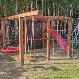 WIKO - Wielofunkcyjny plac zabaw z drewna: huśtawki, ścianka wspinaczkowa z kolorowymi uchwytami, siatka do wspinania i zjeżdżalnia. Dodatkowo widoczny domek na palach ze schodami i zjeżdżalnią w tle...