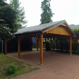 Drewniana konstrukcja z zadaszeniem z dachówki na ceglanym podłożu, widok z zewnątrz, otoczona zielenią i ogrodzeniem z metalowych prętów.