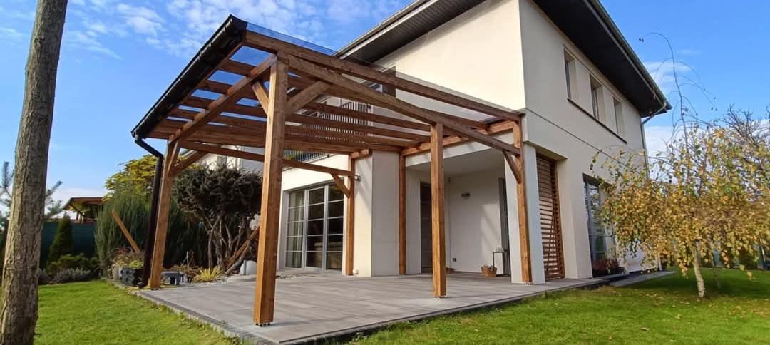 Drewniana pergola z przezroczystym zadaszeniem przylegająca do nowoczesnego domu z jasną elewacją i szarym tarasem, otoczona zielonym trawnikiem i drzewami.